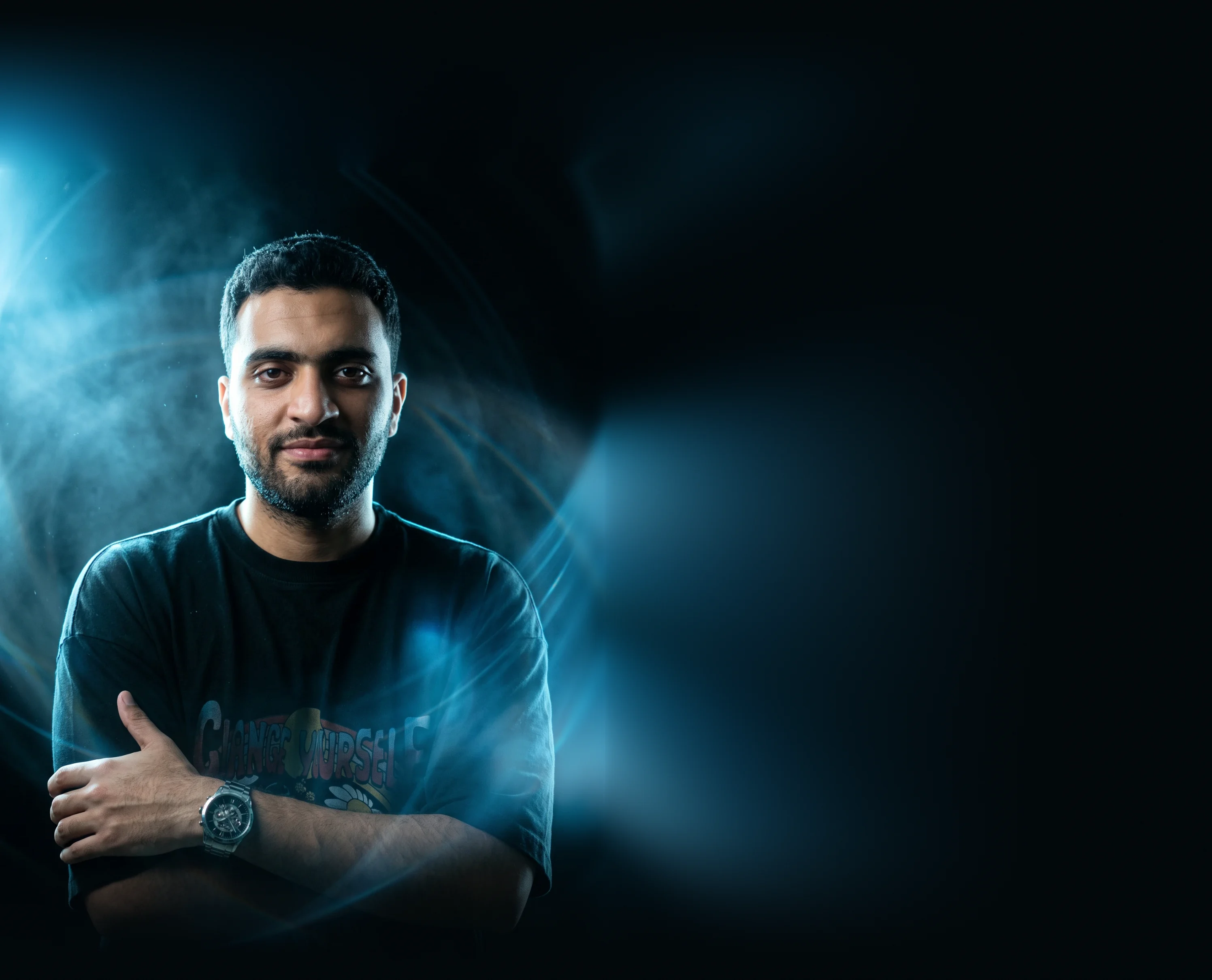 Saleh Elfar smiling slightly with his arms crossed, in a dark T-shirt, with blue light and haze behind him